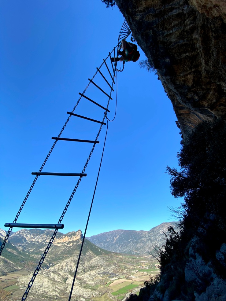 Climber in scaling a twisting via ferrata ladder with the pre-pyrenees in the background
