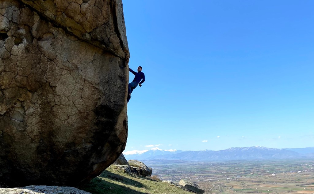 Climber on a highball boulder chalking one hand. There are some flat planes, then with some snowy mountains in the background