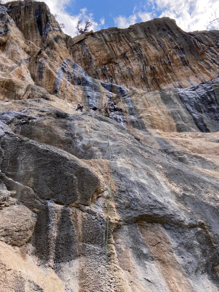 A climber rethreads the rope through an anchor, halfway up a large cliff with many tufa formations and coloured streaks. 