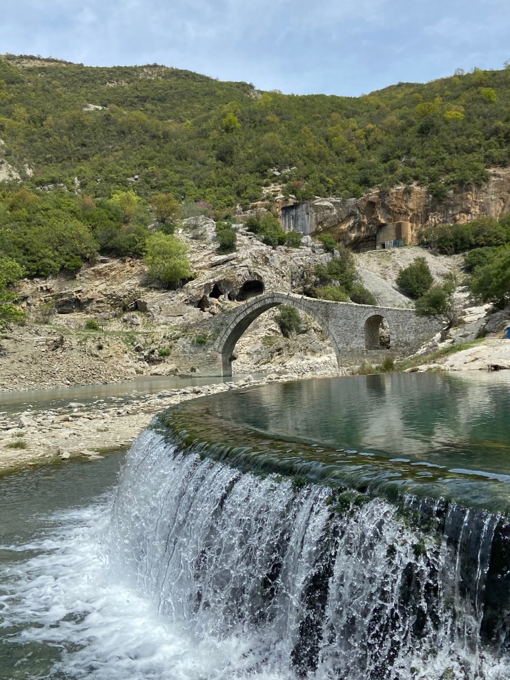 Ottoman style bridge in the background with the thermal spring pool in the foreground. The water is clear and is pouring over the edge of the walls of the pool. There is a small craggy outcrop in the background set upon the trees. 