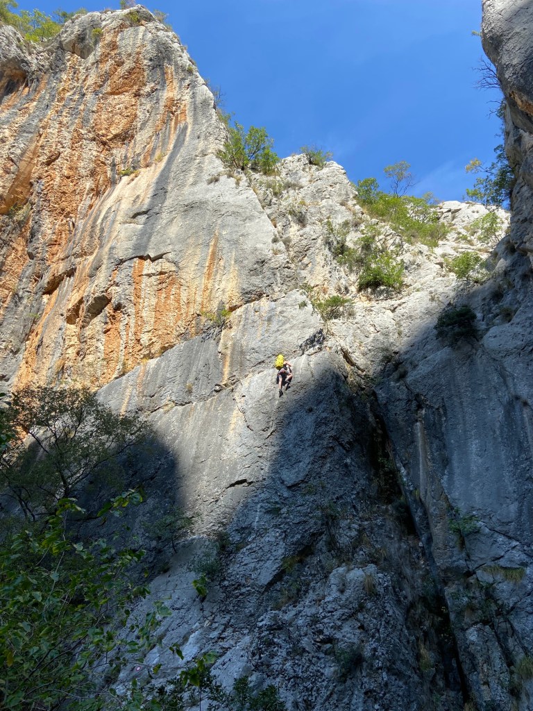 A climber with a large green rucksack ascends a steep section off via ferrata up a cliff. 