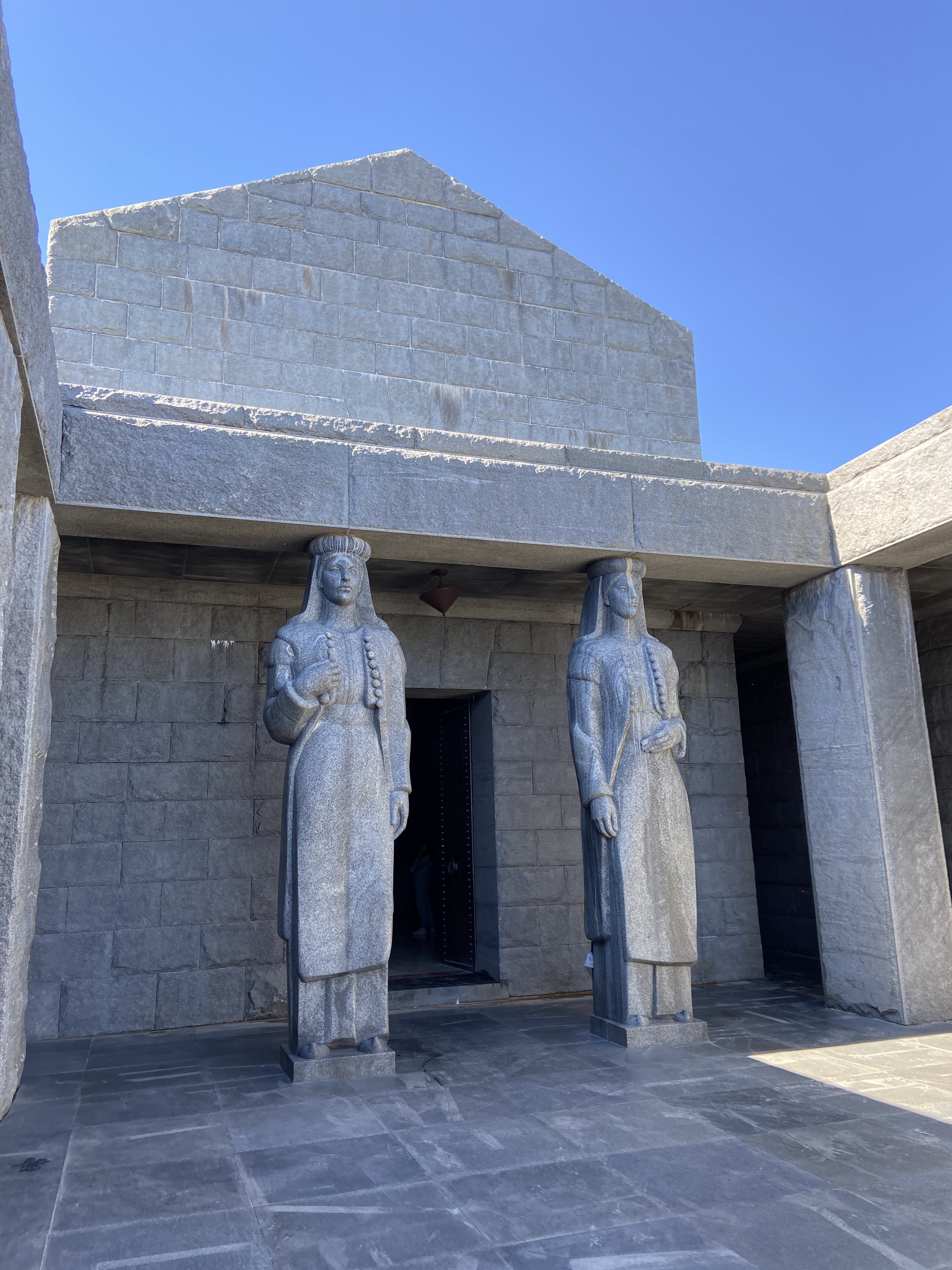 Two giant stone statues stand forebodingly above a mausoleum entrance, whilst also acting as pillars to support the structure. 