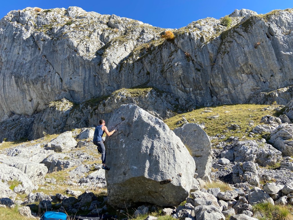 A climber in a blue vest climbs a cool and unique egg shaped limestone boulder, with large mountain cliffs in the background. 