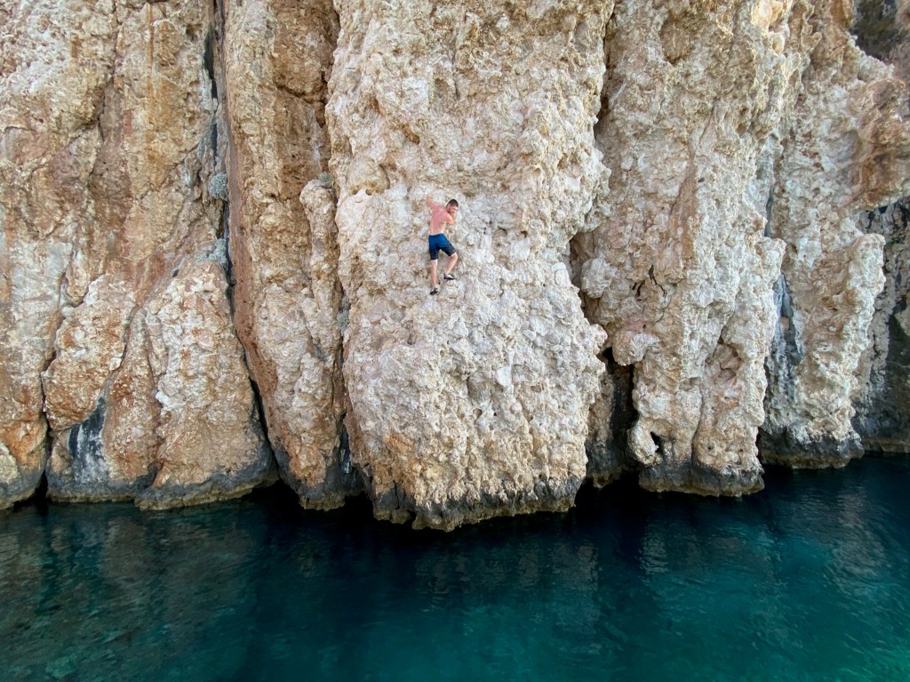 Climber deep water soloing on a white limestone cliff. Around 8 meters above the deep blue sea.