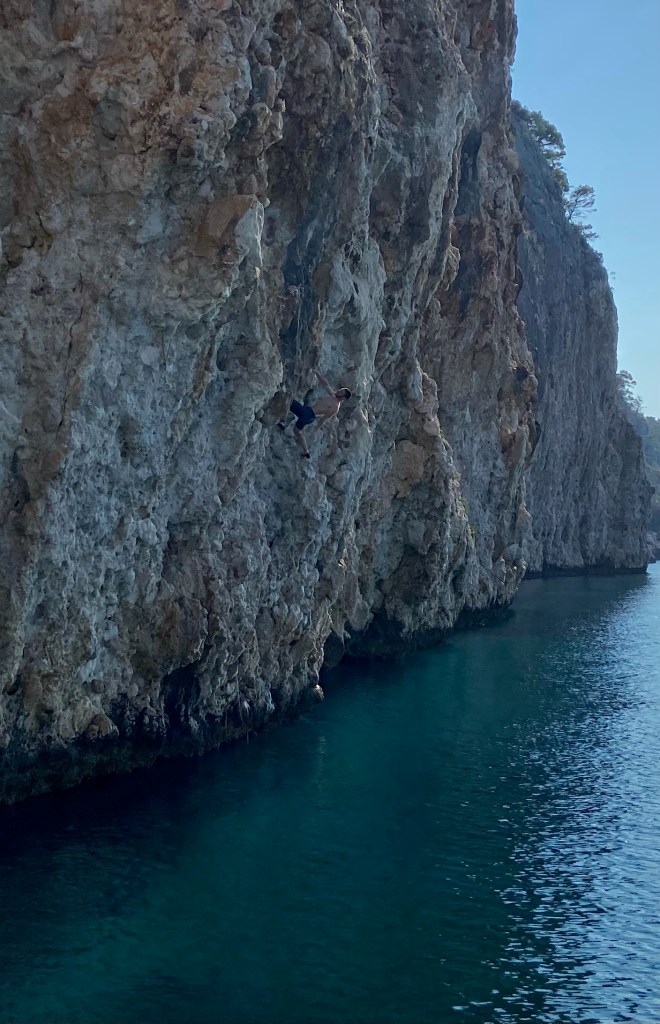 Climber deep water soloing on a white limestone cliff. Around 8 meters above the deep blue sea. The climber has one arm off the wall, resting, and considering their next move.