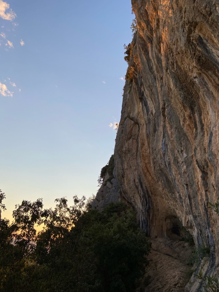 Slightly overhanging cliff dripping in grey and orange tufas. In the background is the sunsetting.