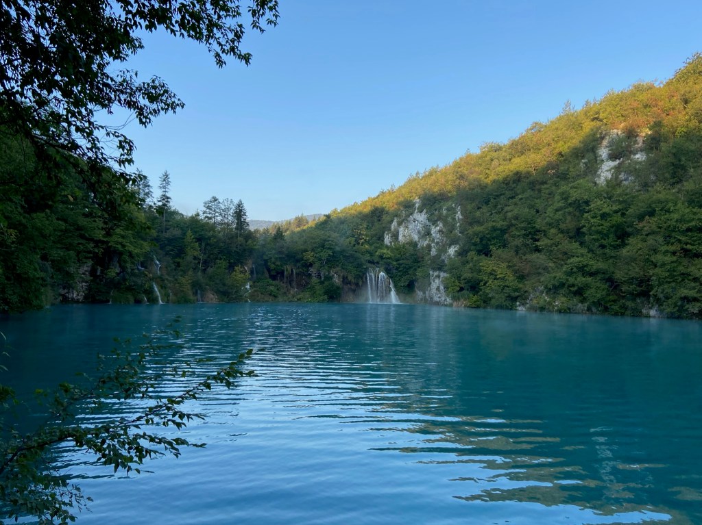 Large blue lake with casades in the background. The lake is surrounded by green forrest.