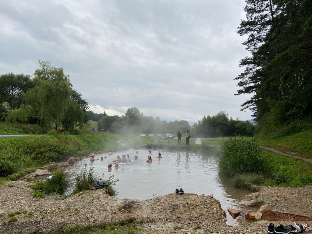People bathing in a pond size body of water which has steam emerging from the surface. There are shoes lined up along the edge and there are cars parked in the field next to the hot spring. 