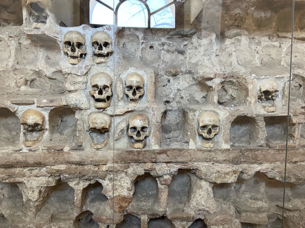 Set behind glass there are human skulls set within recesses of an old stone wall. Most of the recesses are empty as they have been removed over the years.  