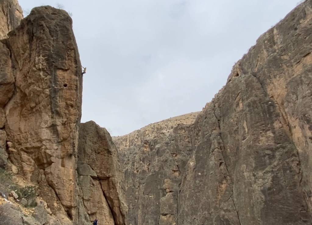 Silhouette of a climber on a pinnacle protruding from the left hand side of the tall sided canyon which is made from dark volcanic rock with long crack feautres.    