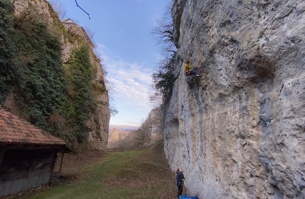 Narrow limestone canyon of Katskhi climbing area. At the bottom there is a wooden tiled hut and grass. On the right hand wall there is a climber on a vertical grey limestone climbing a route. 