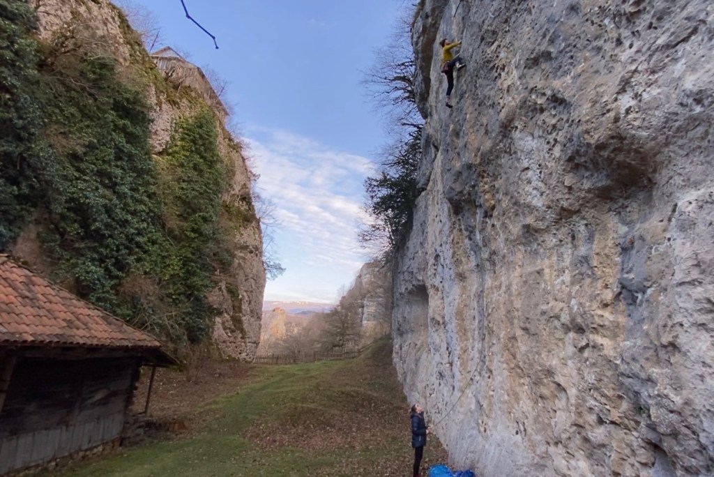 Narrow limestone canyon of Katskhi climbing area. At the bottom there is a wooden tiled hut. On the right hand wall there is a climber on a vertical grey limestone climbing a route.