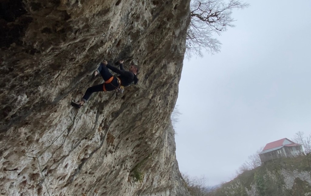 Climber tackling the overhanging section of rock on the short limestone sector. On the other side of the narrow valley is a building set upon another pinnacle. 