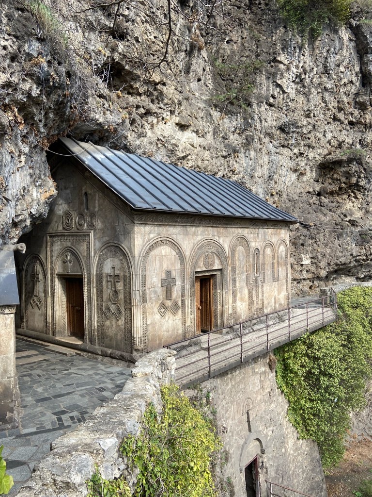Stone church building built into the cliff side. The building has ornate stone carvings including crosses around the two carved wooden doorways