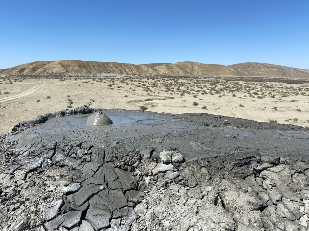 A mud bubble exploding from a dried mud violence. Behind is a desert landscape with a small hill
