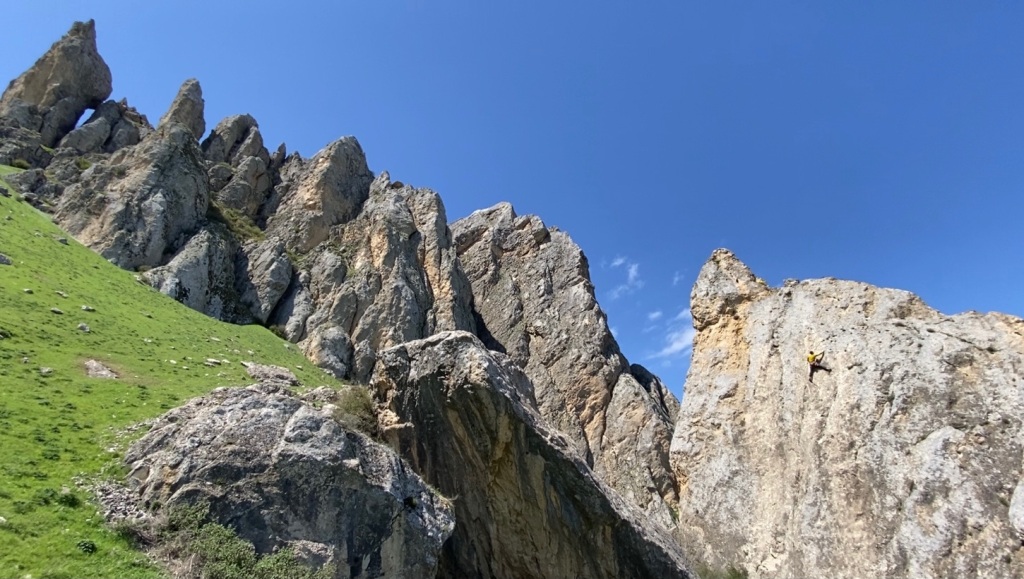 Limestone towers, boulders and pinnacles on a steep grassy hillside with blue sky in the background. There is a climber on one of the boulders