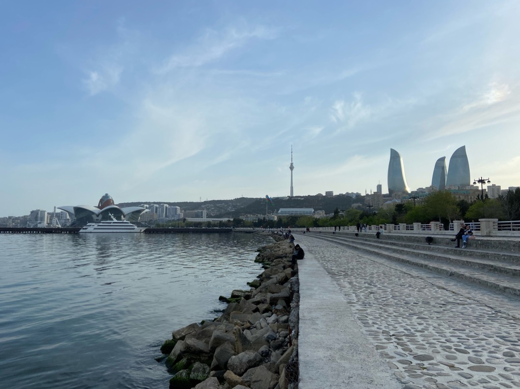 Sea front boulevard with people sitting on step enjoying the surroundings. In the background is Baku flower building where the Eurovision was held, the TV tower and the ionic 3 glass flame towers.
