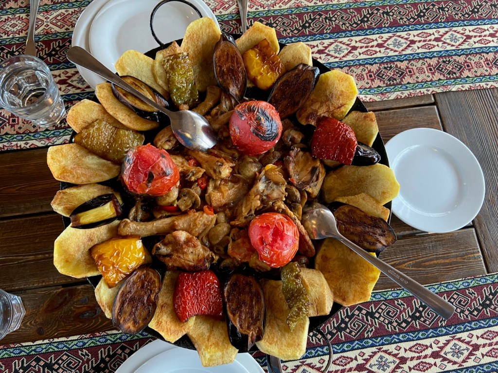Large cast iron plate filled with barbecued chicken in the centre and decorated with grilled peppers, aubergines, tomatoes and potatoes around the edge. 