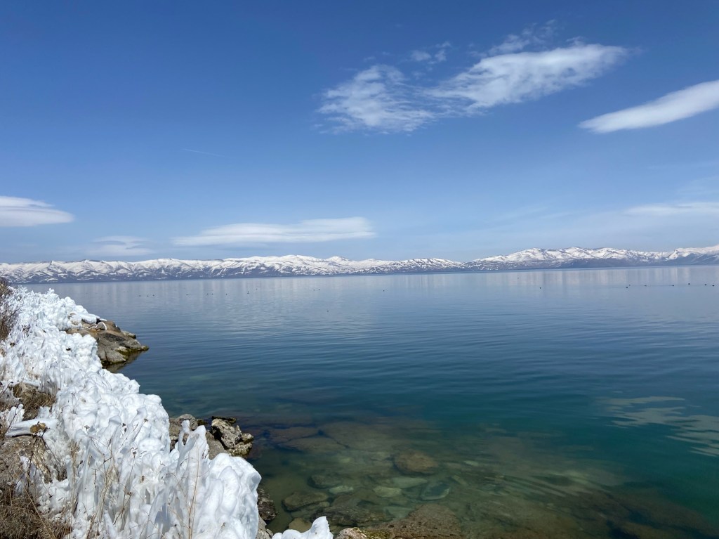 A large alpine lake with snowy peaks in the background. In the foreground are large ice formations on the bank of the lake. 
