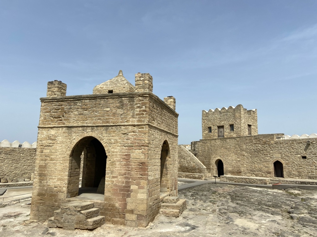 Square building with arches surrounded by a stone wall. Inside the central building there is a flame 