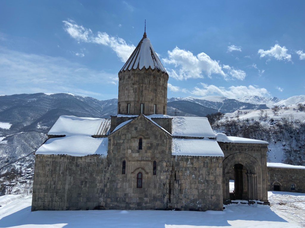 An orthodox monastery covered with snow on the roof and on the ground. There are mountain peaks in the background.