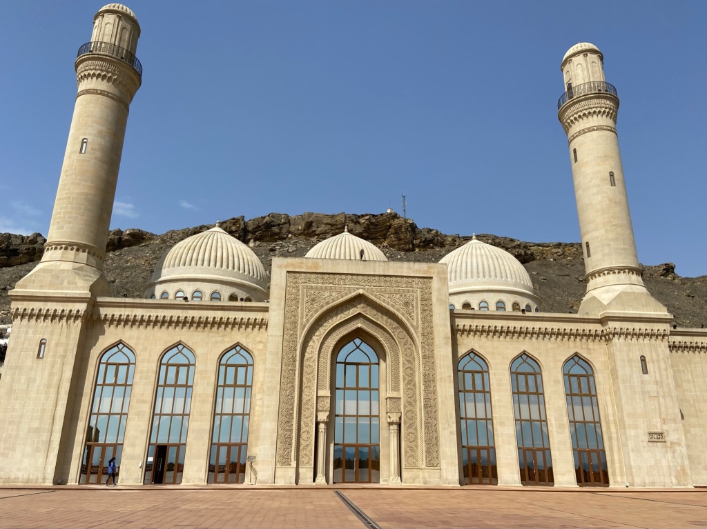 Large and elaborate mosque with two minarets and three large domes. The tall mosque is filled with large windows and the stone has been carved into detailed designs. Behind the mosque is a short rocky crag