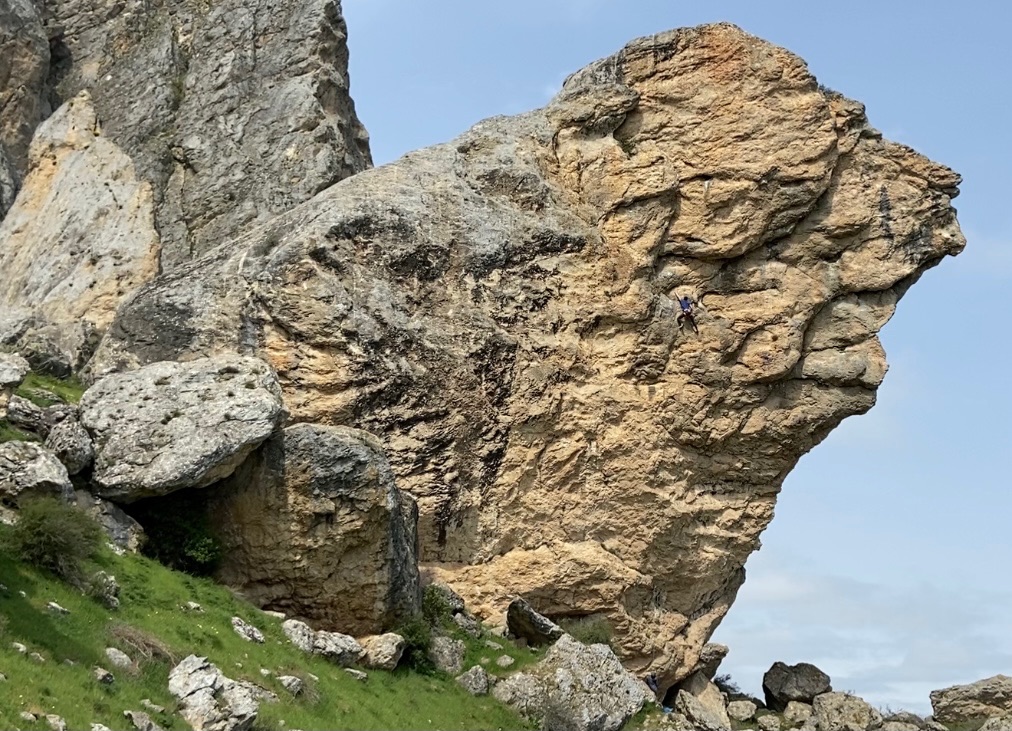 Climber in the middle of a giant pinnacle of rock which is laying on a steep hill side. There is a steep overhanging arete on the right hand side, smaller boulders lying beneath and tall walls of rock in the background. 