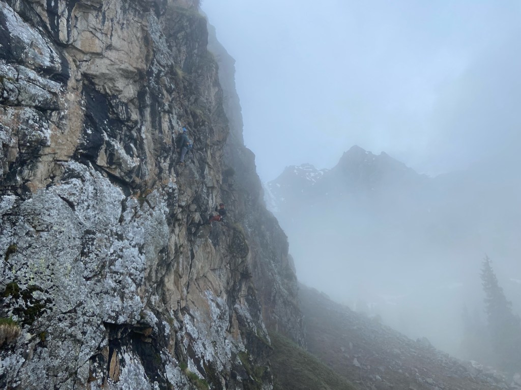 Cliff with climber on. In the background you can see a peak of a taller mountain emerging from the thick fog