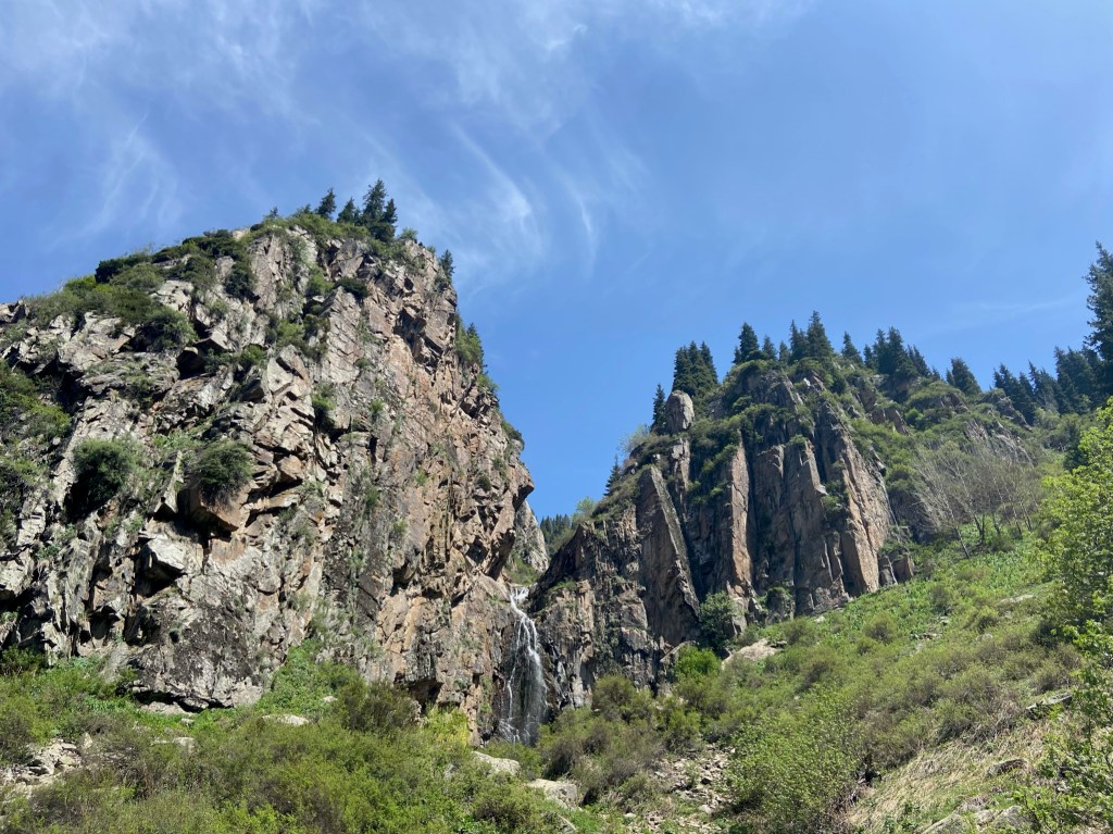 Tall crag with lots of grass ledges and pine trees at the top in the middle there is a waterfall