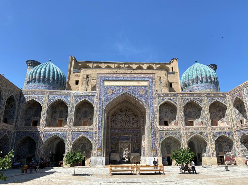Inside one of the Madrasa which has four sides which forms a peaceful country yard. The walls consist of rooms, two stories high, all of which have arches in. The rooms on the ground floor have shops in which are displaying their items for sale like carpets and fabrics. There's two domes on top which are tiled in vibrant blue colours which form intricate patterns. On the front there are also different blue coloured tiles which form swirling patterns. The courtyard has fruit trees and benches in. 