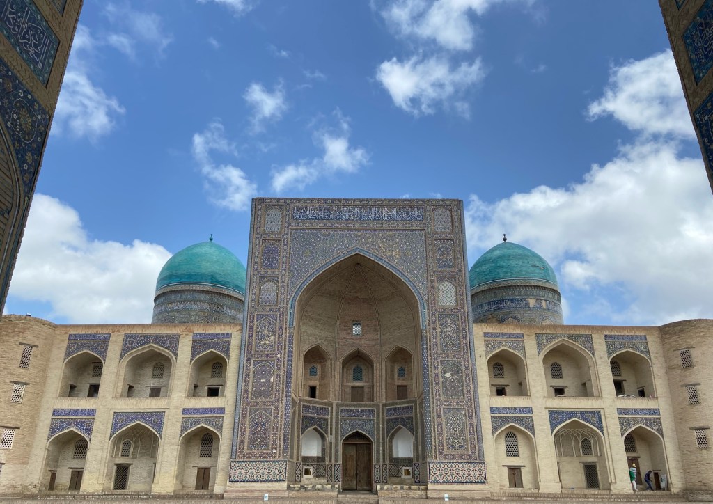 Front one of the Madrasa. There are rooms either side of the tiled entrance. The walls consist of rooms, two stories high, all of which have arches in. There's two domes on top which are tiled in vibrant blue colours which form intricate patterns. 