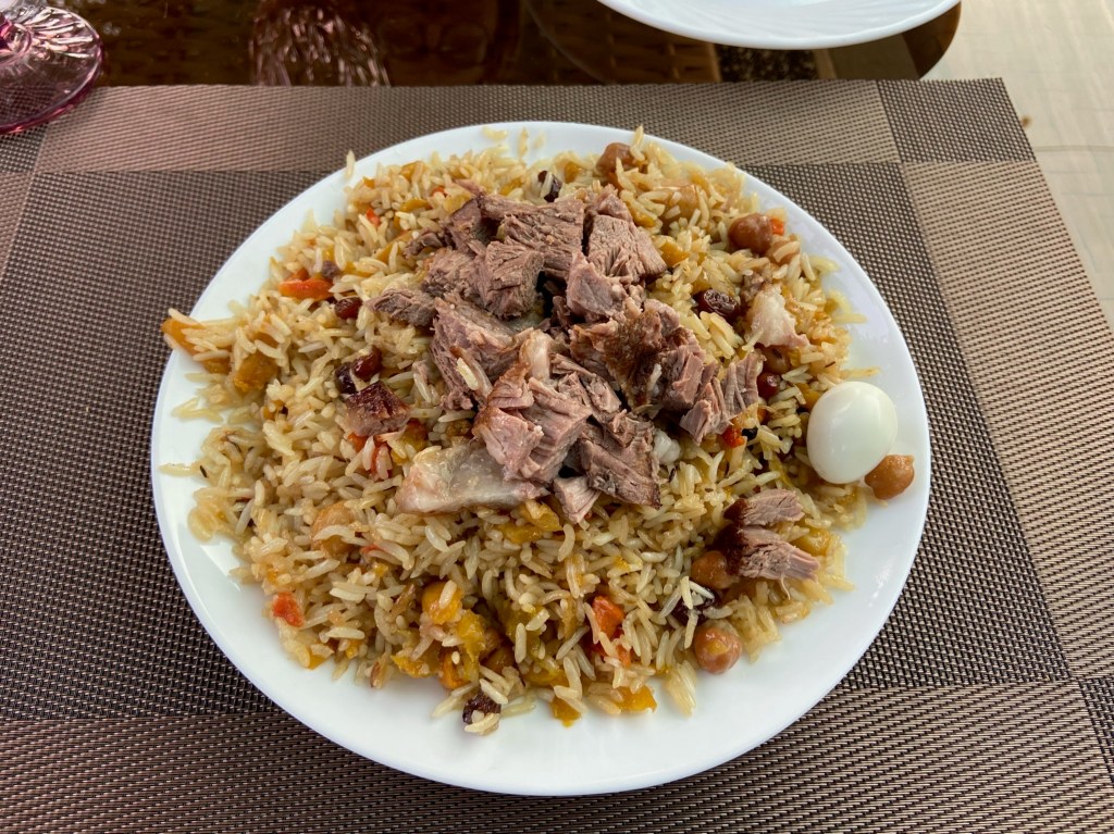 A rice dish which fills the plate. In the rice there are chickpeas, carrot and raisins. On top is a generous portion of lamb and on the side is a small quail's egg.