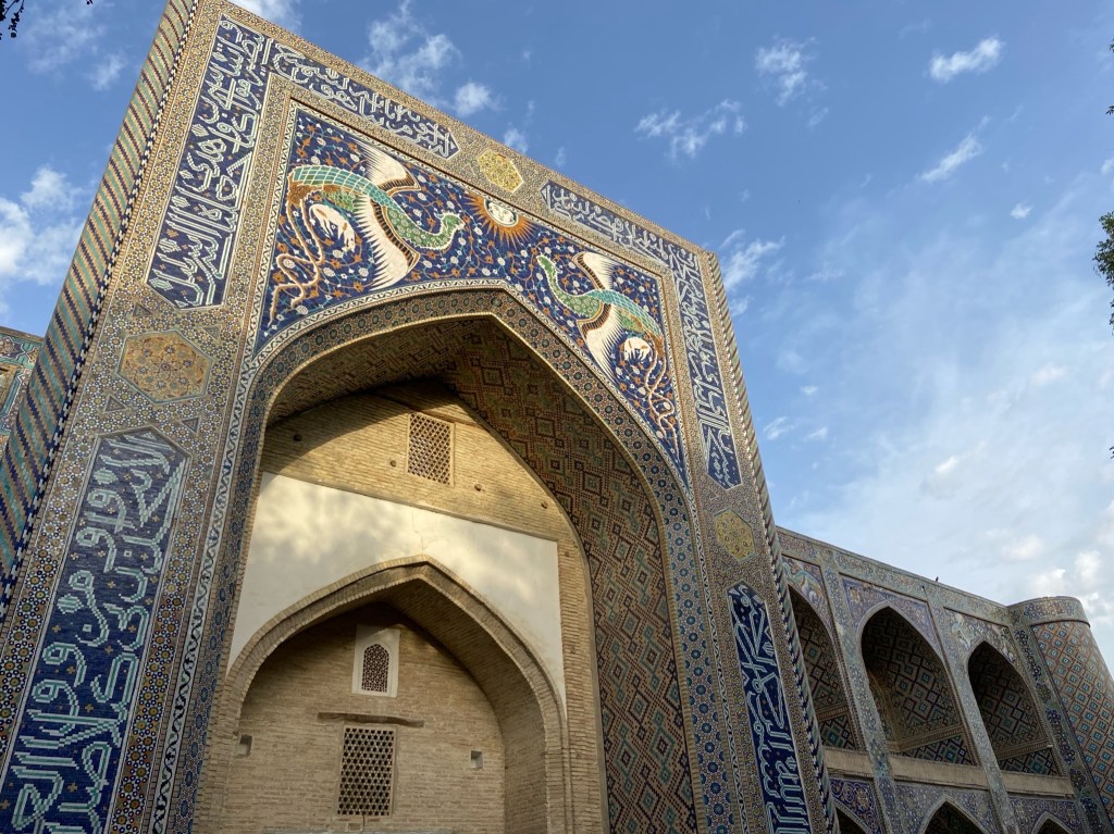A front of a building which is tiled. At the top of the arch entrance is a tiled pattern of two mythical looking birds and a woman's face in the middle of. sun. 
