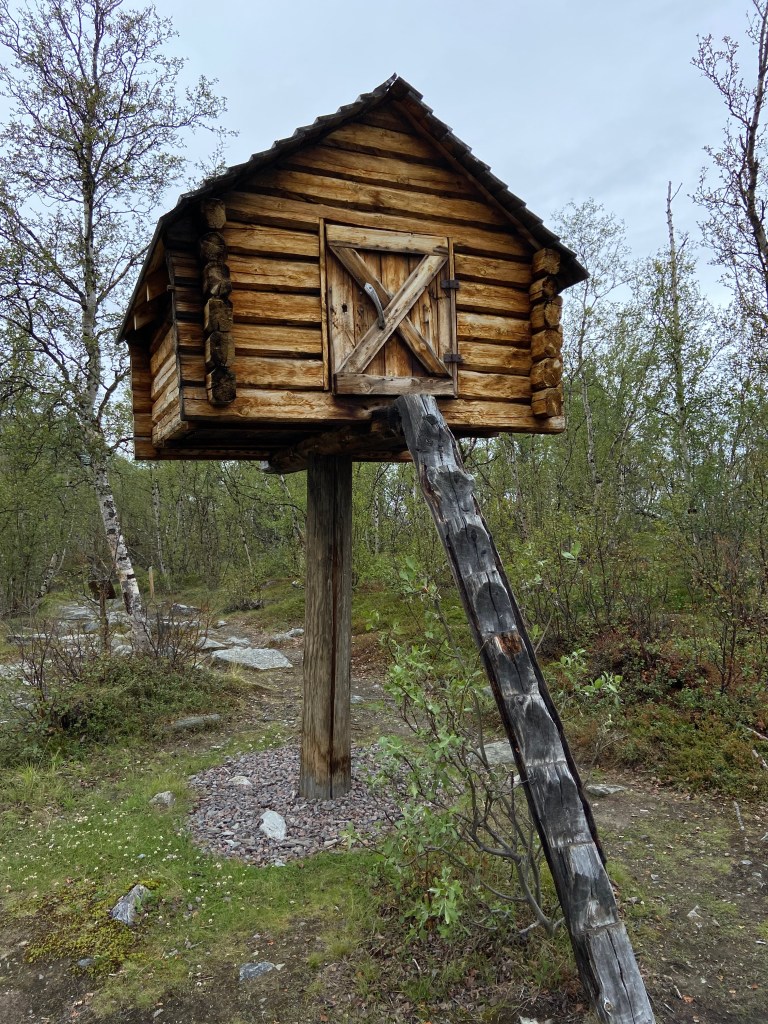 Wooden sami style hut which is balancing on a single pole with another pole allowing access up to the door.
