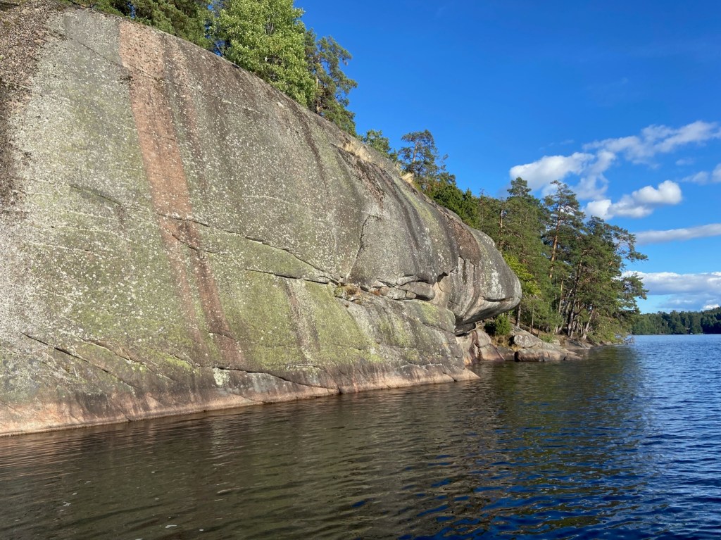 Piece of granite rock with grey and pink streaks which emerges from Lake Ågelsjön. In the background are trees forming a dense forest.