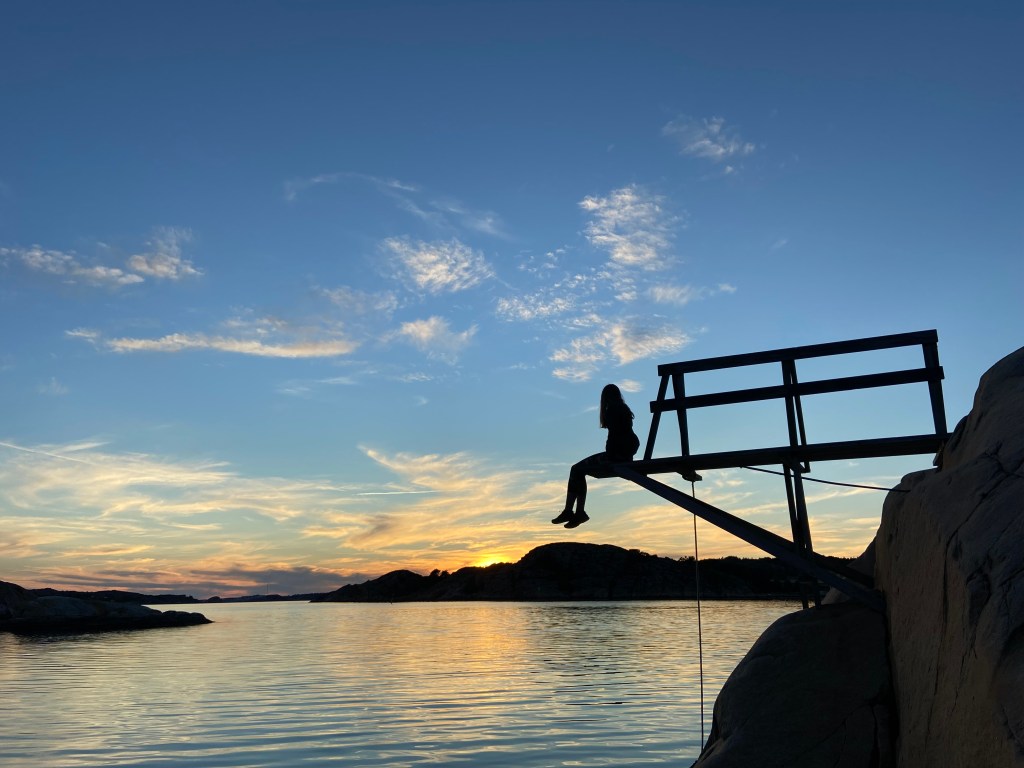 Woman sat silhouetted on the end of a diving board above the sea. The sun is setting behind an island which creates a calm bay.