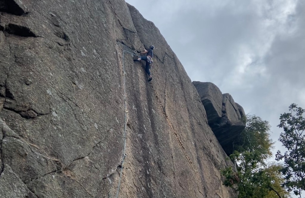 Climber on granite cliff working along the horizontal and vertical cracks. In the crack is trad protection. The sky above is grey and cloudy. 
