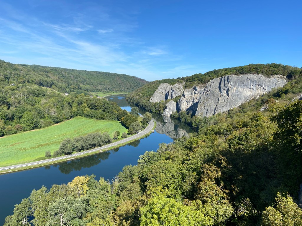 Limestone fins protruding out form the slow moving river. On the other side following the river is a road with green fields and forest. 