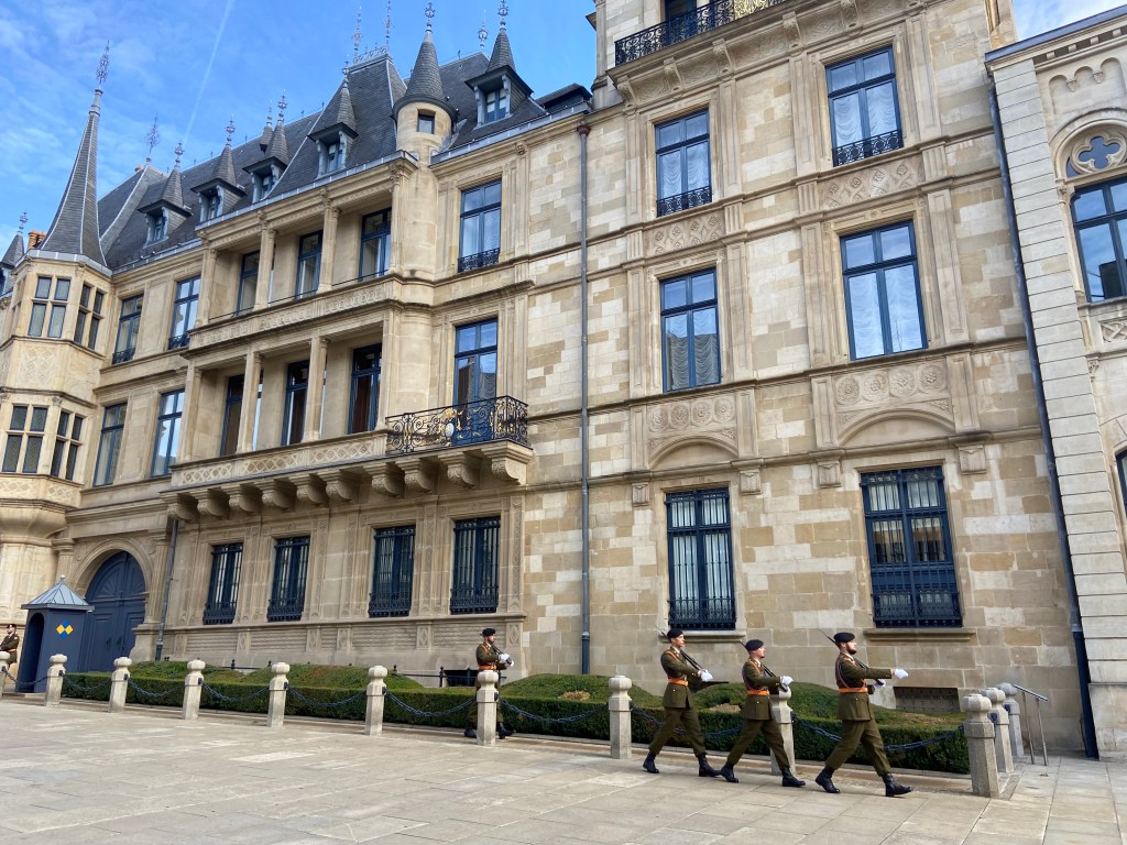 Grand palace building made from light coloured stone. There are many windows and some decorative turrets and balconies. In front there are four military guards parading and keeping guard with guns.