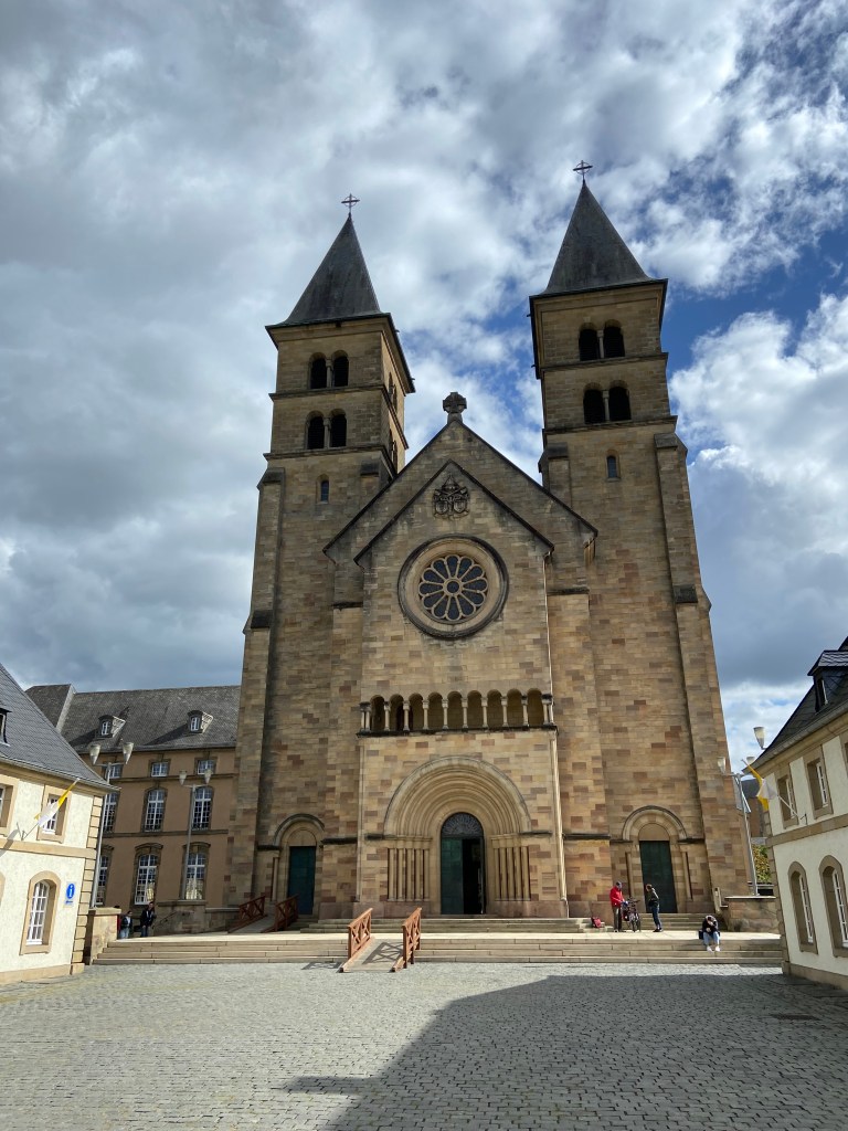 A grand stone abbey with a large circular decorative window and two towers either side. In front of the church is a cobbled courtyard 