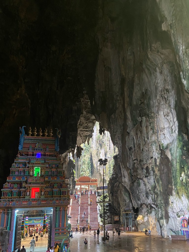 Set inside the cave is a large colourful temple with a door and behind is a set of stairs leading to another temple in a cave with an opening in the ceiling allowing light to flood in.