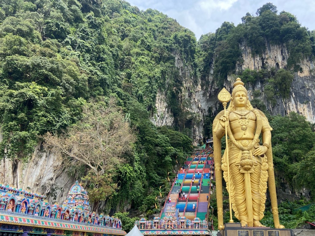 Large golden statue of the Hindu God Murugan. Behind is a colourful and steep stairway which leads up into the side of the hill and into a cave complex.