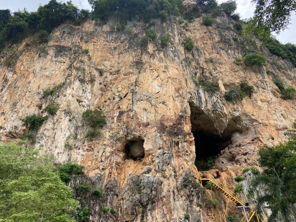 Orange white limestone cliff around 70 metres tall with black strikes. There is a yellow staircase leading up to a cave half way up the cliff. On the left there's a small cave with a massive boulder chockstone jammed in the back.