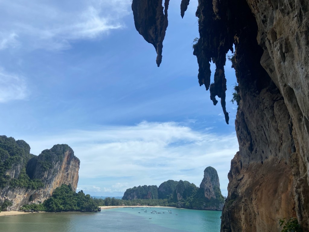 Looking out of a cave which is lined with tufas along the edges. On the opposite side across the bays are Tonsai and Railay. There are two beaches with cliffs on each side.