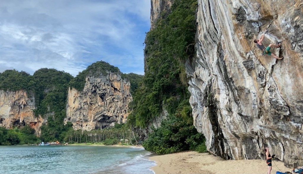Climber lay backing on the curved flake of Lion King. The belayer on Ton Sai beach . On the opposite side of the bay are more limestone cliffs with caves and tufas surrounded by jungle.