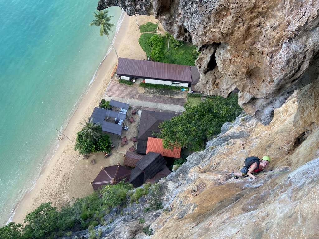 Climber on the exposed crux pitch of Humanality with a huge tufa hanging behind. Below there is a bar on the beach and the blue sea. 