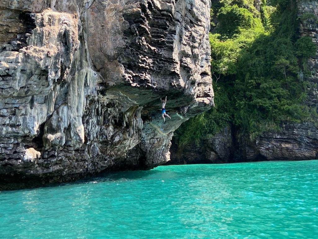 Climber deep water soloing, hanging from only their arms on a steep overhanging roof section of limestone high above the turquoise blue sea. 