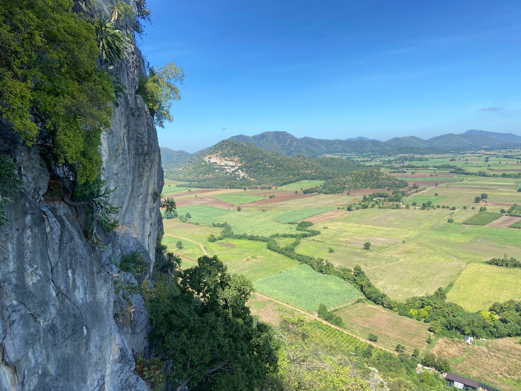Tall grey limestone cliff surrounded by green fields with some smaller tree covered mountains in the background 