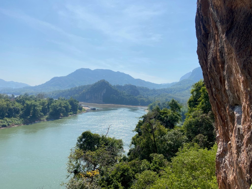 Orange limestone wall towering over the slow wide river on the opposite side of the river are forested hills and blue sky