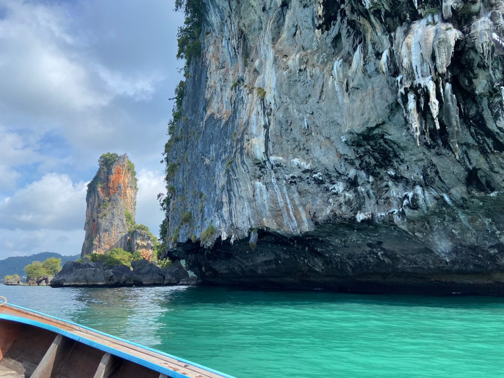 Boat alongside a tall grey limestone cliff with a steep under cut bottom which is dripping with tufas. In the background is the tall orange and grey of Ao Nang Tower standing alone in the sea